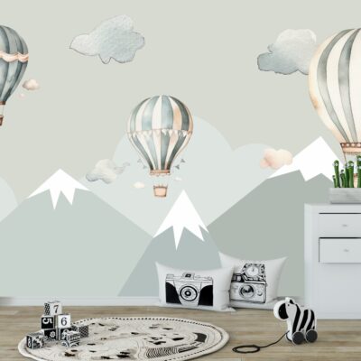Fototapeta ścienna dla dzieci przedstawiająca góry ośnieżone szczyty w odcieniach szarości i błękitu, nad którymi unoszą się trzy retro balony na ogrzane powietrze w pastelowych barwach: szaro-miętowe i beżowe. Wokół balonów widoczne są delikatne, akwarelowe chmurki. Na pierwszym planie widoczne są elementy wystroju dziecięcego pokoju: duża, wzorzysta litera "A", czarno-białe klocki z cyframi, okrągły dywanik z motywem drogi, poduszki z grafikami aparatów fotograficznych, zebra na kółkach oraz biała komoda z rośliną w doniczce i pluszowymi zabawkami.
