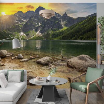 Wizualizacja nowoczesnego salonu z dużą fototapetą na całej tylnej ścianie, przedstawiającą górski krajobraz z jeziorem, przypominający Morskie Oko w Tatrach, oświetlony zachodzącym słońcem. Po lewej stronie pokoju stoi biała sofa z poduszkami przy dużym oknie, a w centrum okrągły, szary stolik kawowy na geometrycznej podstawie, stojący na szarym dywanie. Nad stolikiem wisi beżowa, designerska lampa. Po prawej stronie znajduje się zielony fotel na drewnianych nogach oraz wysoki, industrialny regał z roślinami i książkami. Podłoga wykonana jest z ciemnego drewna.