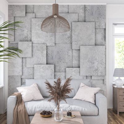 Jasna aranżacja salonu w stylu nowoczesnym, w której centralną ścianę zdobi szara fototapeta imitująca trójwymiarowe, betonowe bloki i kwadraty o różnej wielkości i głębokości. Na tle ściany stoi jasnoszara, dwuosobowa sofa z dwiema jasnoróżowymi poduszkami i przewieszonym brązowym pledem. Przed kanapą znajduje się niski, drewniany stolik kawowy, na którym stoi szklany wazon z bukietem suszonych traw ozdobnych. Nad strefą wypoczynkową wisi brązowa, ażurowa lampa w stylu boho. Po lewej stronie widać duże okno oraz fragment rozłożystej, zielonej rośliny doniczkowej. Po prawej stronie znajduje się drugie okno, a pod nim drewniana komoda z szufladami, na której stoi mała biała lampka. Podłoga wyłożona jest ciemnymi panelami drewnianymi.