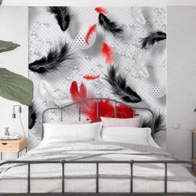 Aranżacja nowoczesnej sypialni z łóżkiem o czarnej, metalowej ramie w stylu industrialnym. Ścianę za wezgłowiem zdobi wyrazista fototapeta z motywem spadających piór. Pióra są przedstawione w trójwymiarowej stylistyce; część z nich jest czarna, część biała (z delikatną fakturą), a kompozycję ożywiają jaskrawe, czerwone pióra stanowiące mocny akcent kolorystyczny na jasnym tle. Pościel jest biała, z jedną poduszką w kolorze pudrowego różu. Po lewej stronie widać dużą zieloną roślinę doniczkową, a po prawej niską, drewnianą szafkę z dekoracjami.