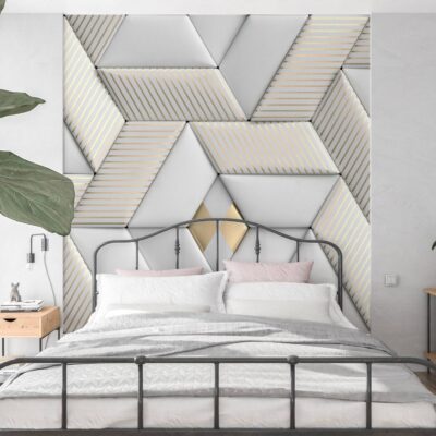 Aranżacja przytulnej sypialni z łóżkiem o czarnej, metalowej ramie w stylu retro. Ściana za wezgłowiem ozdobiona jest fototapetą z geometrycznym wzorem 3D, przypominającym układankę z trójkątów i trapezów. Grafika utrzymana jest w jasnej kolorystyce bieli, kremowego beżu i szarości, przełamanej eleganckimi, złotymi akcentami. Niektóre elementy wzoru imitują miękką strukturę materiału, inne gładkie powierzchnie. Po lewej stronie kadru widoczne są duże liście rośliny doniczkowej, a po prawej niska biała komoda z dekoracjami i książkami.