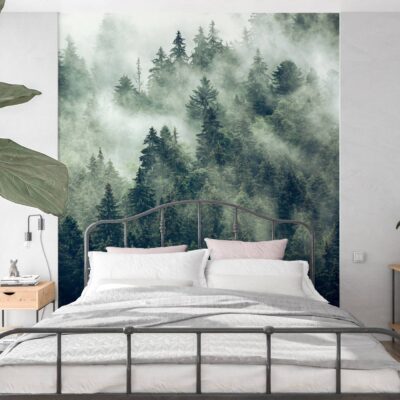 Zdjęcie sypialni z dużym łóżkiem o czarnej, metalowej ramie. Całą ścianę za zagłówkiem zajmuje fototapeta z widokiem na las iglasty. Czubki sosen i świerków wyłaniają się z gęstej, mlecznobiałej mgły, co tworzy atmosferę spokoju i tajemniczości. Wnętrze jest jasne, z drewnianymi akcentami i żywymi roślinami doniczkowymi po bokach.