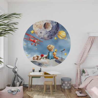 Aranżacja pokoju dziecięcego z dużą okrągłą naklejką tkaninową Mały Książę i Lis. Granatowa akwarelowa ilustracja z czerwonym samolotem, planetami i różą umieszczona nad drewnianym biurkiem, obok łóżka z różowym baldachimem.