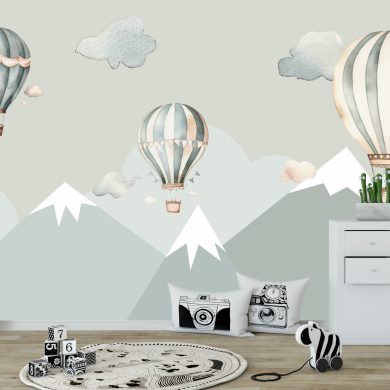 Fototapeta ścienna dla dzieci przedstawiająca góry ośnieżone szczyty w odcieniach szarości i błękitu, nad którymi unoszą się trzy retro balony na ogrzane powietrze w pastelowych barwach: szaro-miętowe i beżowe. Wokół balonów widoczne są delikatne, akwarelowe chmurki. Na pierwszym planie widoczne są elementy wystroju dziecięcego pokoju: duża, wzorzysta litera "A", czarno-białe klocki z cyframi, okrągły dywanik z motywem drogi, poduszki z grafikami aparatów fotograficznych, zebra na kółkach oraz biała komoda z rośliną w doniczce i pluszowymi zabawkami.